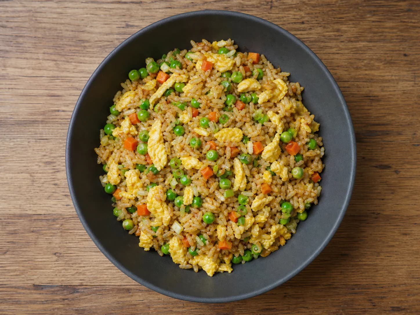 Vegetable fried rice with egg ribbons, peas, carrots and scallions in a dark bowl.